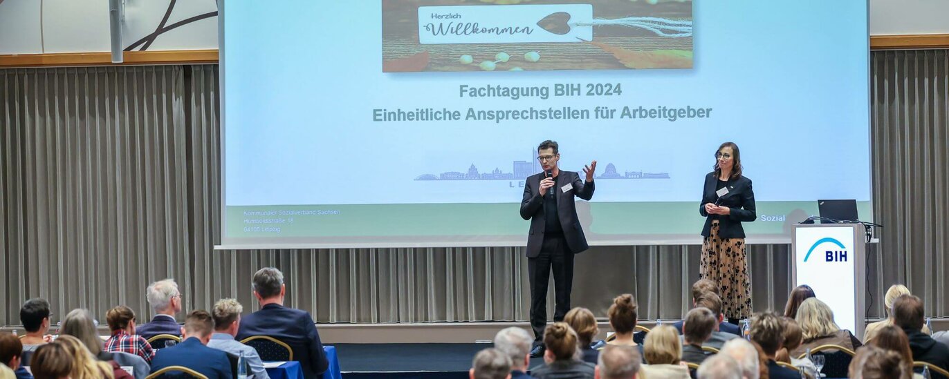 Das Foto zeigt einen Mann (links) und eine Frau (rechts) auf der Bühne eines großen Saales. Auf der Leinwand steht: Herzlich Willkommen! BIH Fachtagung 2024 Einheitliche Ansprechstellen für Arbeitgeber