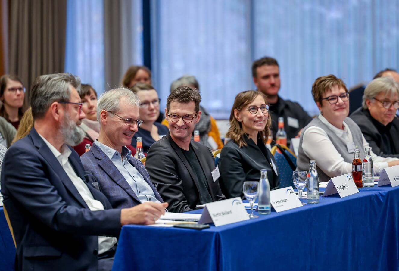 Das Foto zeigt einen Saal mit Teilnehmenden. In der vorderen Reihe sitzen drei Männer und zwei Frauen - sie lächeln.