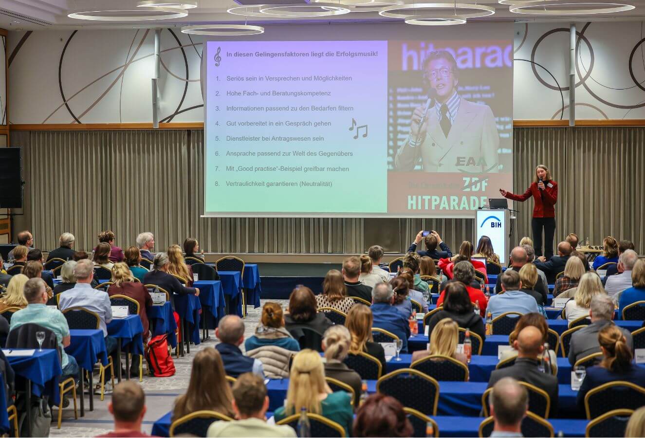 Das Foto zeigt einen großen vollbesetzten Saal mit langen Stuhlreihen. Der Blick fällt auf die Leinwand auf der Bühne. Dort stehen verschiedene Punkte unter der Überschrift: "Hitparade des Gelingens"