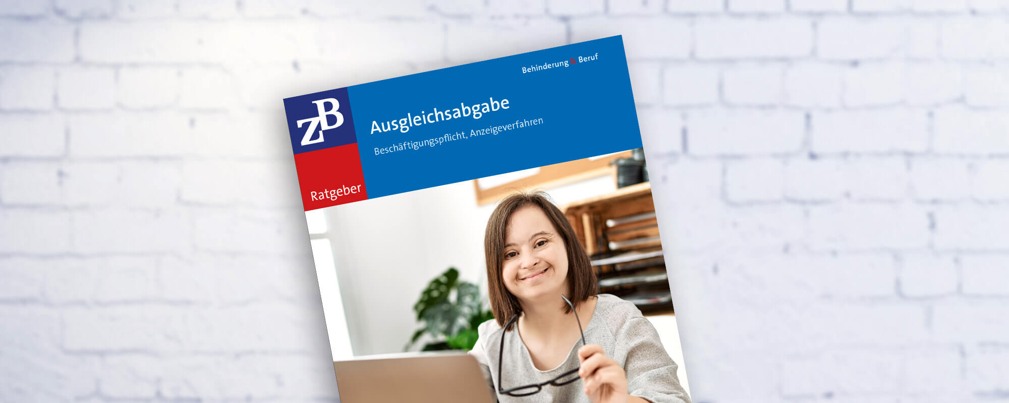 Die Fotomontage zeigt das Cover der Zeitschrift ZB Ratgeber Ausgleichsabgabe vor einer weißen Ziegelwand. Auf dem Cover ist eine Frau mit Downsyndrom an einem Computerarbeitsplatz zu sehen.