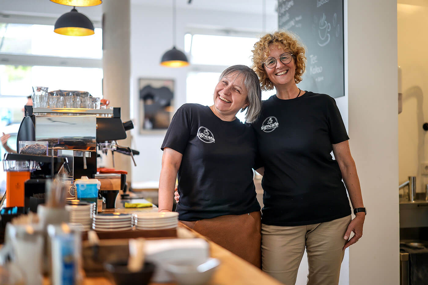 Zwei Frauen stehen Arm in Arm rechts von der Theke eines Cafés. Die Geschäftspartnerin legen lächelnd die Köpfe aneinander..