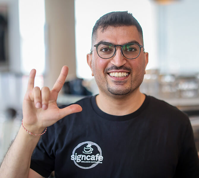 Ein junger Mann in einem schwarzen T-Shirt und dem weißen Aufdruck "signcafe. Inklusive Kaffeebude"formt mit der rechten Hand die Gebärde für "I love you".