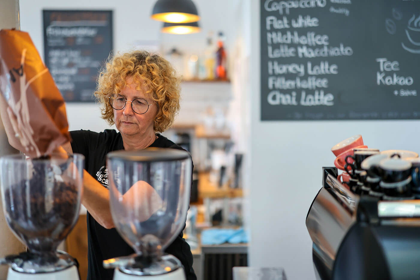 Eine Frau mit rotblonden Locken und schwarzem T-Shirt füllt den Kolben einer großen Kaffeemaschine mit Bohnen auf.