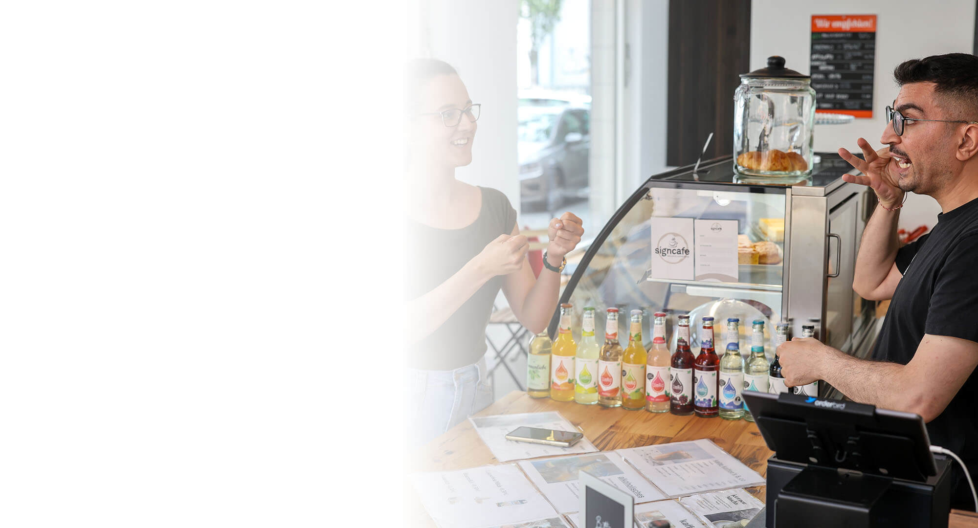 Ein junger Mann mit schwarzem T-Shirt und dunklen Haaren steht hinter der Theke eines Cafés und unterhält sich in Gebärdensprache mit einer Kundin.