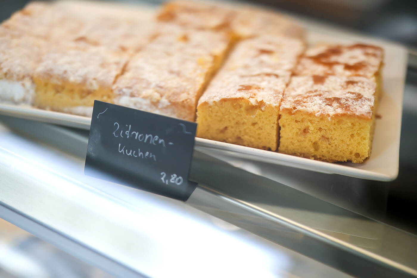 In der Kuchentheke eines Kaffees steht eine weiße längliche Platte mit Zitronenblechkuchen.