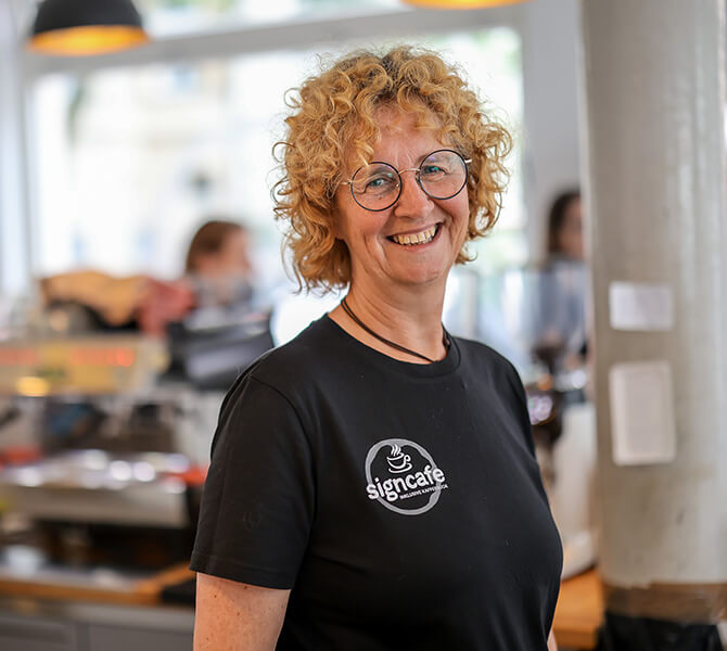 Eine Frau mittleren Alters mit rotblonden Locken und schwarzem T-Shirt lächelt direkt in die Kamera. Auf dem T-Shirt steht "signcafe. Inklusive Kaffeebude"