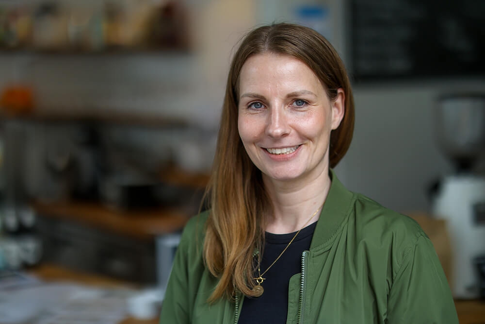 Eine junge Frau mit langen Haaren und grüner Blouson-Jacke steht lächelnd in einem Café.