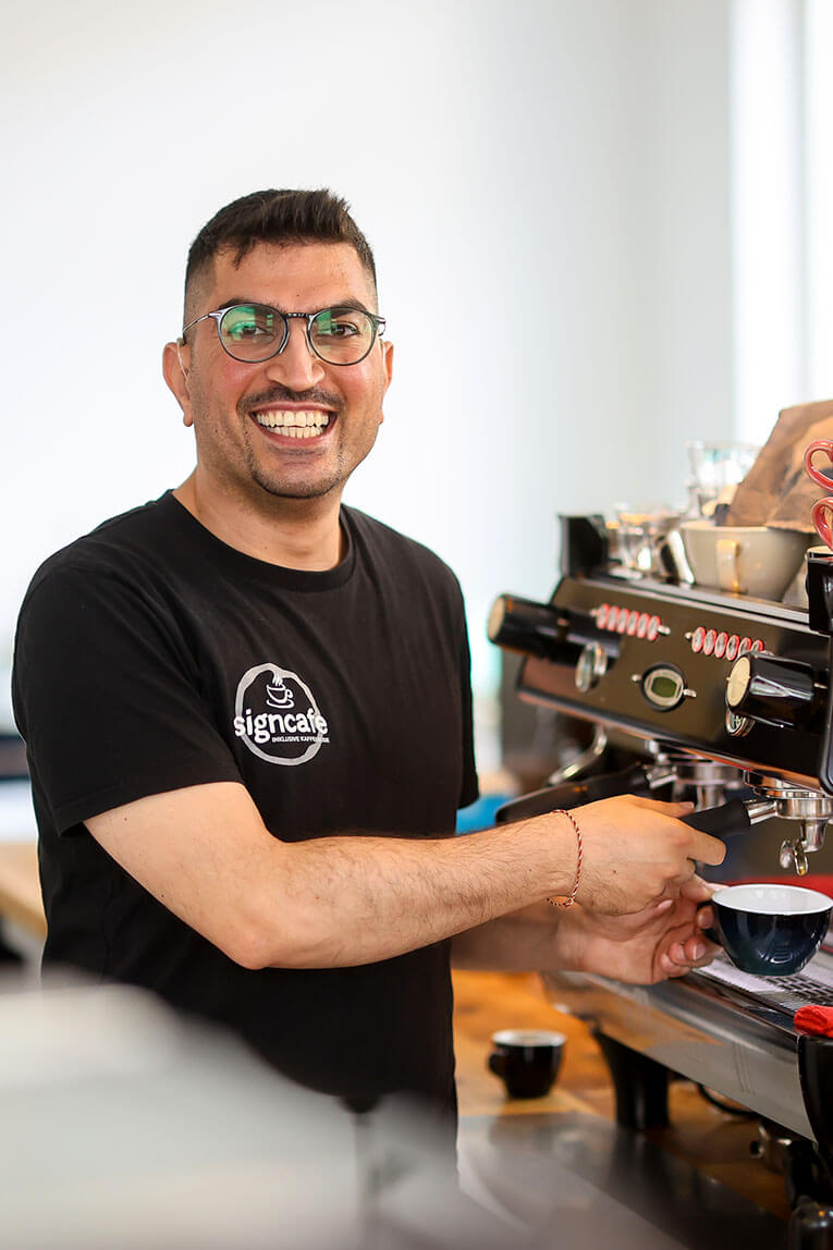 Ein junger Mann steht links von einer großen Siebträgerkaffeemaschine. Mit der rechten Hand Hand schiebt er eine blaue Tasse unter den Siebträger. 