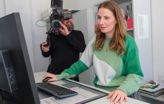 Eine junge Frau steht an einem Stehschreibtisch und arbeitet an einem PC. Hinter ihr steht ein Kameramann und filmt sie dabei.