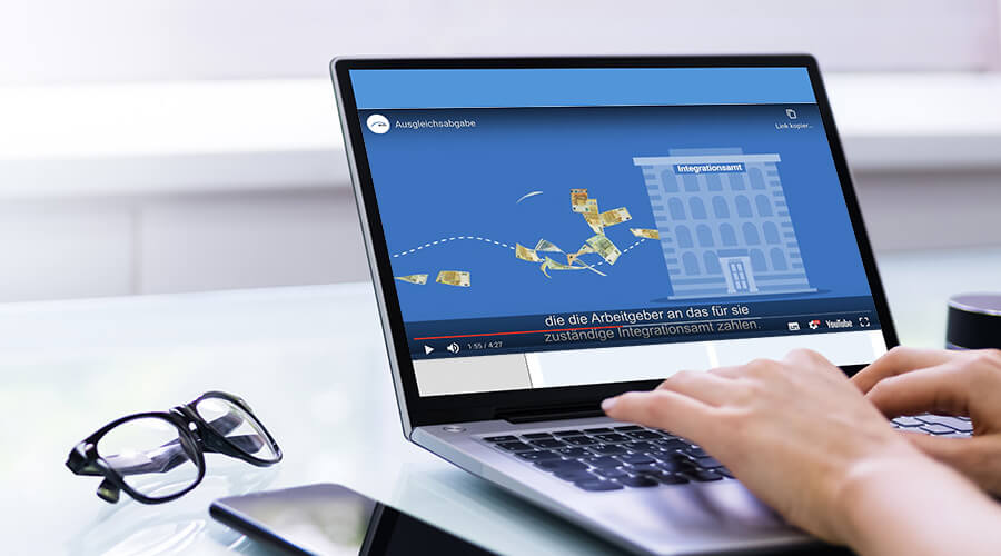 Auf einem Laptop-Monitor ist ein Ausschnitt eines Erklärfilms zur Ausgleichsabgabe zu sehen. In der Szene fliegen Geldscheine in ein Gebäude, das die Inklusions- und Integrationsämter symbolisiert.