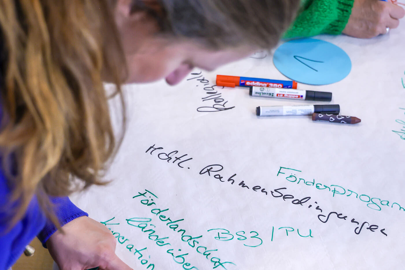 Eine Frau beugt sich über ein großes Blatt Papier, auf dem sie mit bunten Filzschreibern schreibt.