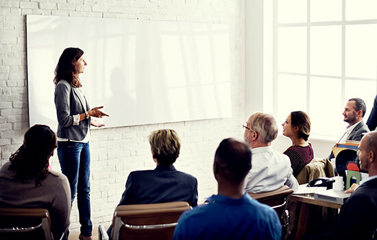 Eine Frau präsentiert vor einer Gruppe in einem hellen Raum mit Whiteboard und großen Fenstern. Die Teilnehmenden sitzen in Business-Kleidung und hören aufmerksam zu.