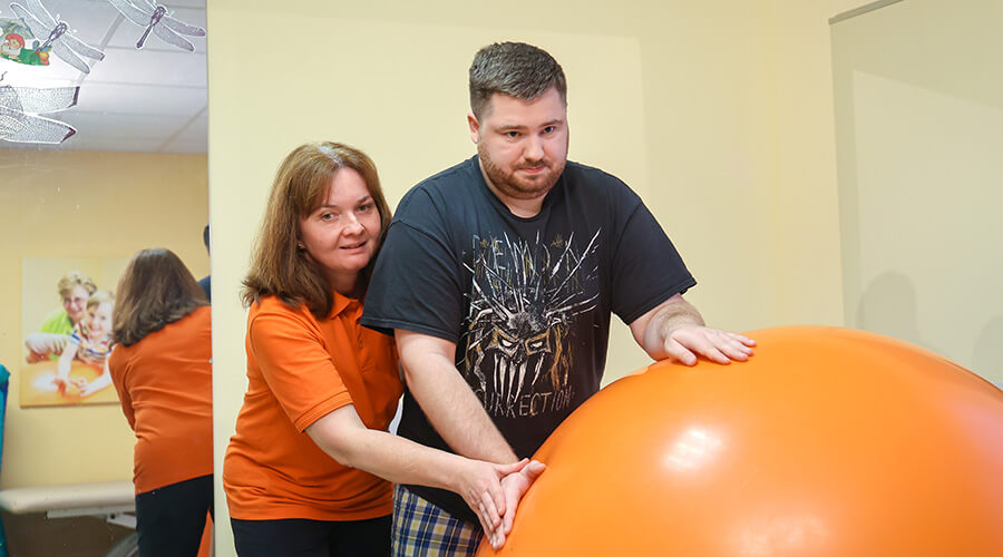 Eine Physiotherapeutin behandelt einen jungen Mann, der sich auf einen großen Gymnastikball abstützt. Die Therapeutin steht hinter ihm.