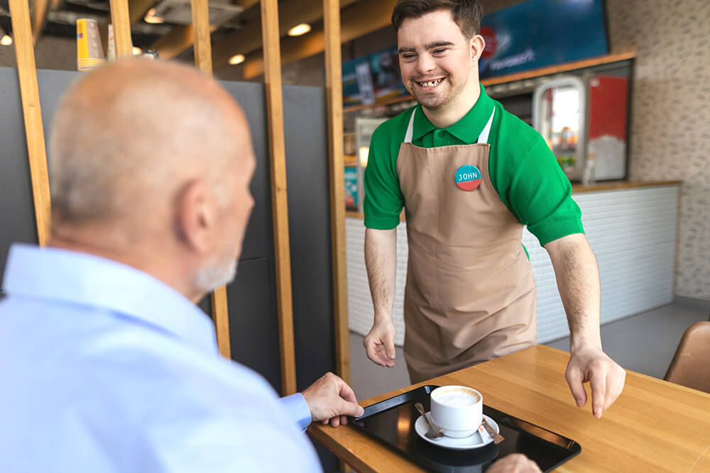 Ein junger Mann mit Down-Syndrom serviert einen Kaffee, er lacht. 