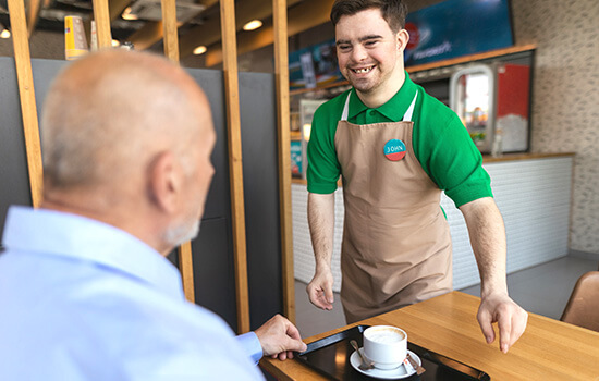 Ein junger Mann mit Down-Syndrom serviert einen Kaffee. Er lacht.