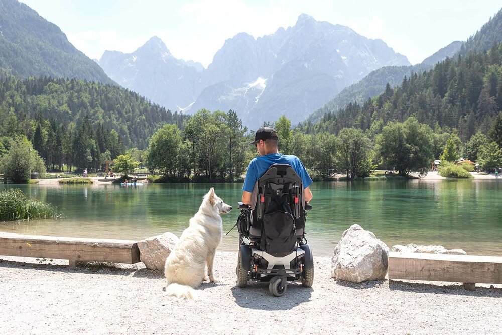 Ein Mann im Rollstuhl sitzt am Ufer eines Sees, neben ihm sitzt ein weißer Hund. Der Mann sitzt mit dem Rücken zur Kamera. Im Hintergrund ist ein Bergpanorama zu sehen,